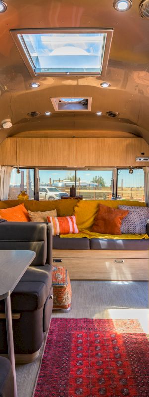 The image shows a cozy, modern RV interior with a seating area, kitchen, and colorful accents, well-lit with sunlight and stylish decor.