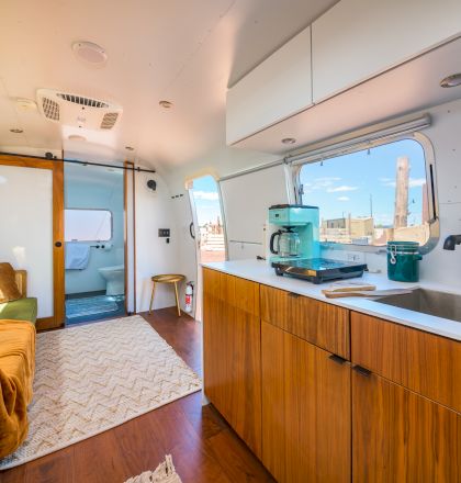 Modern RV interior with wooden cabinets, cozy seating, a rug, and kitchen appliances, bathed in sunlight from the window.
