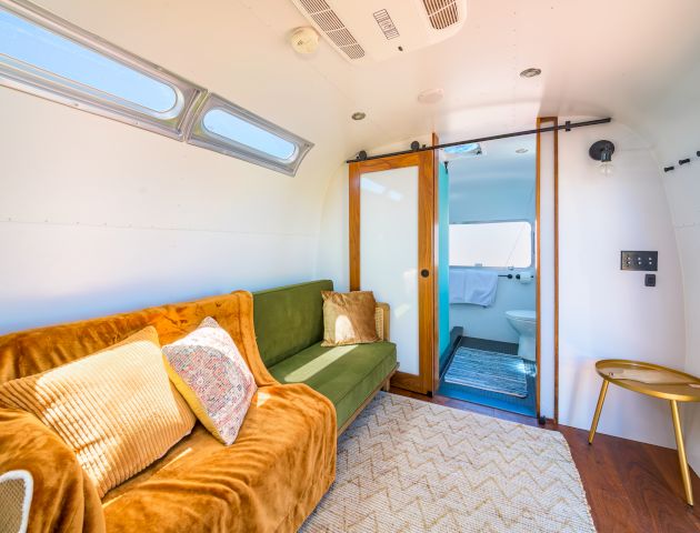 The image shows a modern tiny home interior with a green sofa, a sliding door leading to a bathroom, cozy decor, and a bright atmosphere.