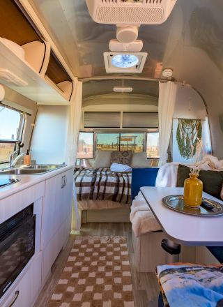 The image shows a cozy modern camper interior with a small kitchen, dining area, and bed, featuring bright natural lighting and stylish decor.