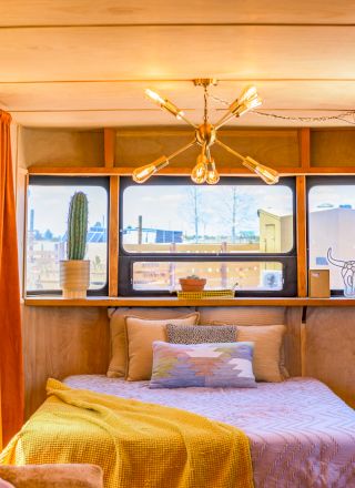 Cozy bedroom with a bed, colorful bedding, and large windows. Decor includes warm lighting, orange curtains, and cactus decor items.