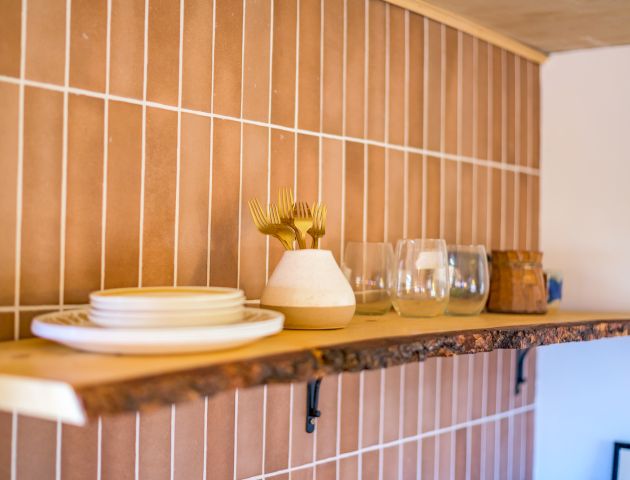 A wooden shelf with plates, a vase, cutlery, glasses, and a small container against a tiled wall.